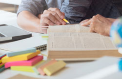 Duas mãos apontando para trechos de um livro aberto em cima de uma mesa, com blocos adesivos coloridos e cadernos espalhados.