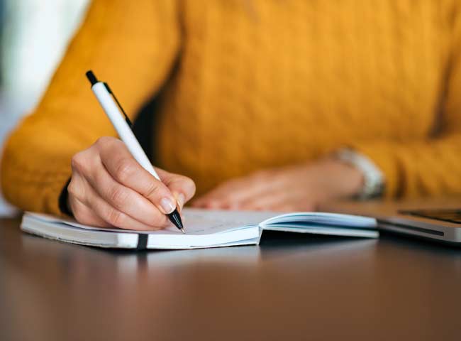 Imagem de uma estudante vestindo uma blusa de lã laranja escrevendo em seu caderno com uma caneta branca e preta.