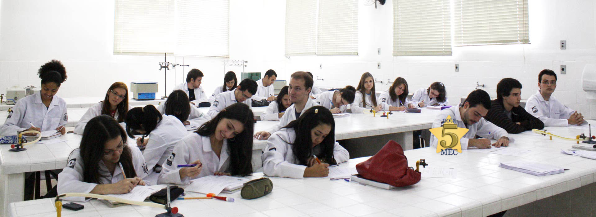 Diversos alunos vestidos de jalecos brancos sentados escrevendo.