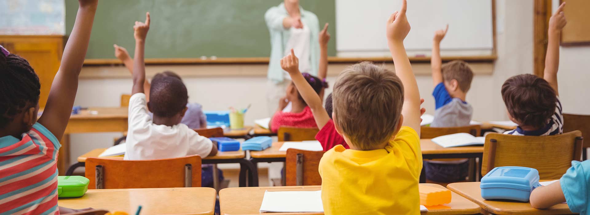 Vista de trás de crianças sentadas em uma sala de aula com as mãos levantadas.