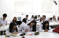 Diversos alunos vestidos de jalecos brancos sentados escrevendo.