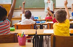 Vista de trás de crianças sentadas em uma sala de aula com as mãos levantadas.